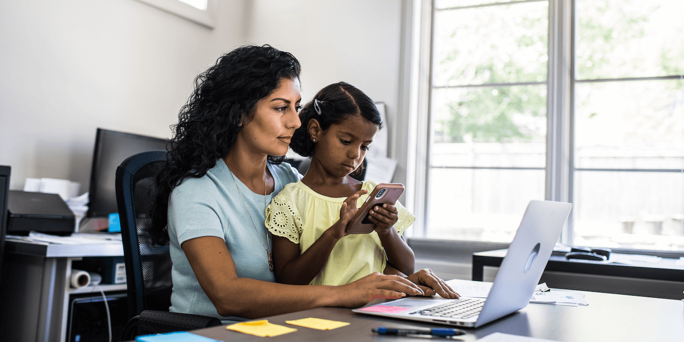 parent working on a laptop while their child sits in their lap and looks at a cell phone