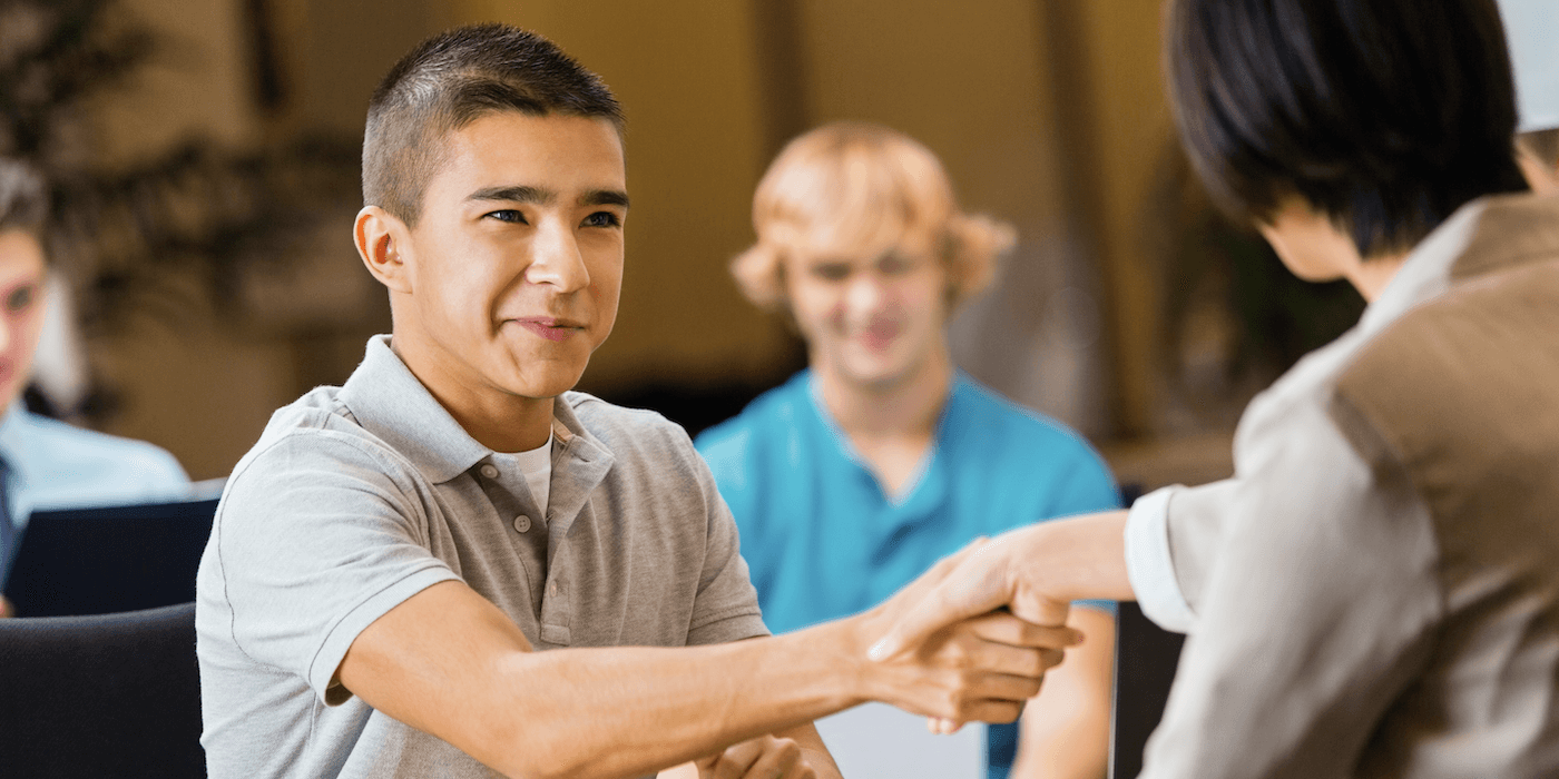 high school student shaking hands with an interviewer at a job fair with at least one other student visible in the background
