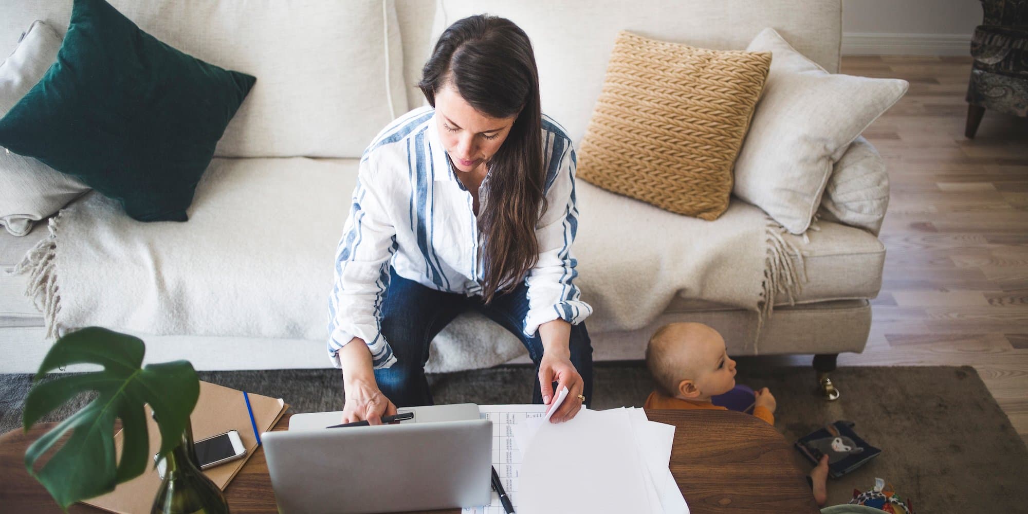 person sitting on couch, working at coffee table looking at papers and laptop while toddler sits on the floor