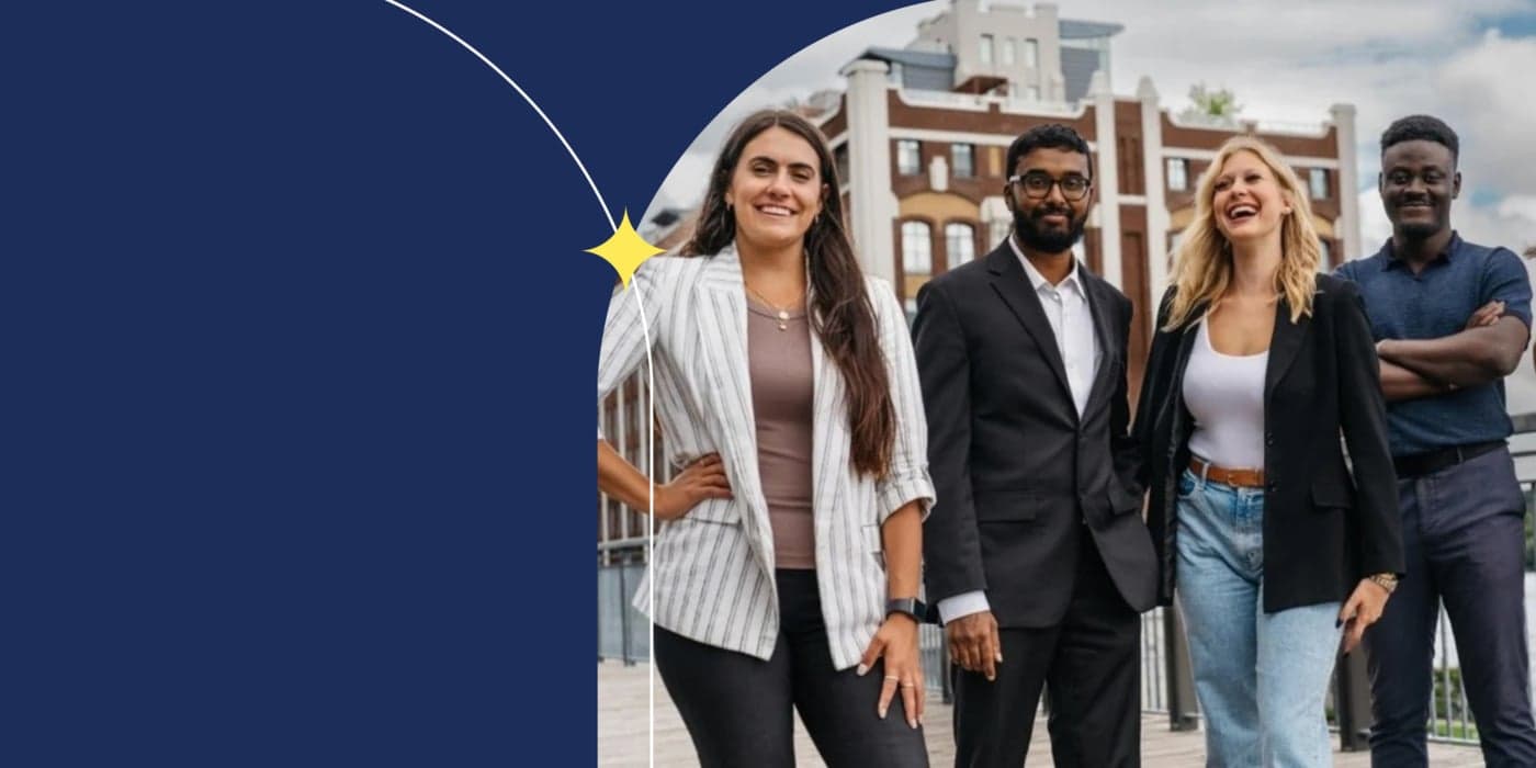 Four people are standing in front of a historic-looking brick building, smiling and laughing. The person on the left side of the photo is wearing a white blazer with grey stripes. They have long dark hair. The person next to them is wearing a black suit with a white collared shirt. They have short curly dark hair. The person in the middle has on a black blazer and a white scoop-neck. They have shoulder-length blonde hair and are laughing. The person on the right is wearing a navy polo shirt and has dark, curly hair. They have their arms folded across their chest.