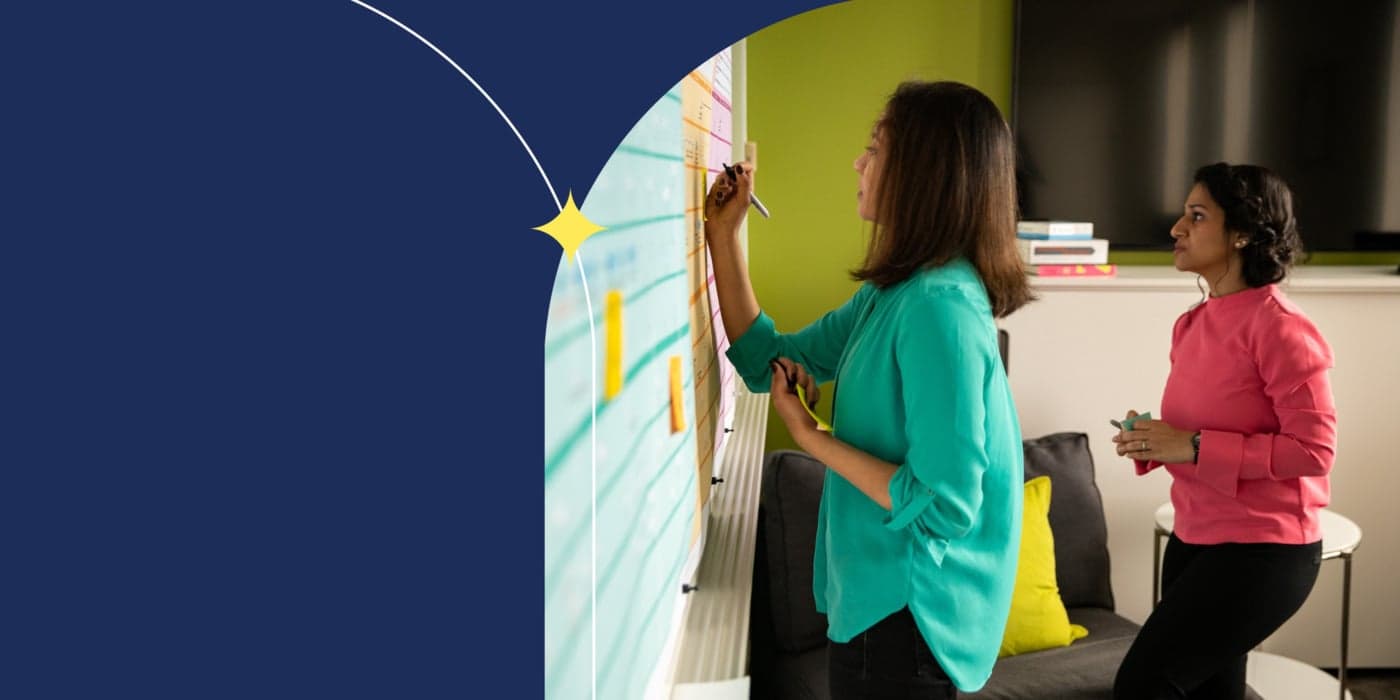 Two people standing in front of a whiteboard with focused looks on their faces. The one standing at the board writing has shoulder-length dark brown hair and is wearing a teal blouse. She is holding a Sharpie marker. The person standing behind her has dark brown French braids and is wearing a magenta blouse.