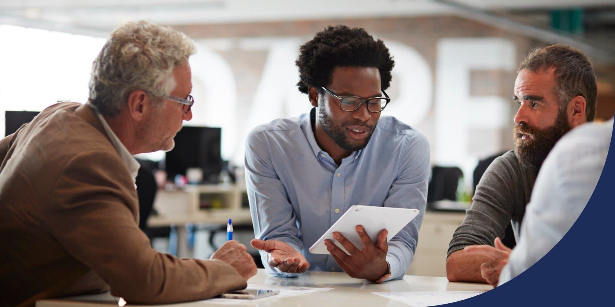 Black professional sitting in an office, holding a tablet with one hand and the other palm turned upward, and talking to white coworkers